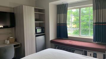 Habitación de hotel con cama, escritorio con computadora, microondas y refrigerador pequeño. Se ve una ventana con cortinas y vistas a árboles desde el exterior.