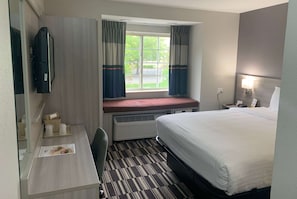 A hotel room with a bed, a desk with a chair, a TV mounted on the wall, a window with curtains, and a nightstand with a lamp. - Microtel Inn by Wyndham Charlotte Airport (Charlotte)