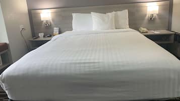 Cama bem arrumada com roupa branca, mesa de cabeceira com abajur, telefone e um aviso na parede.