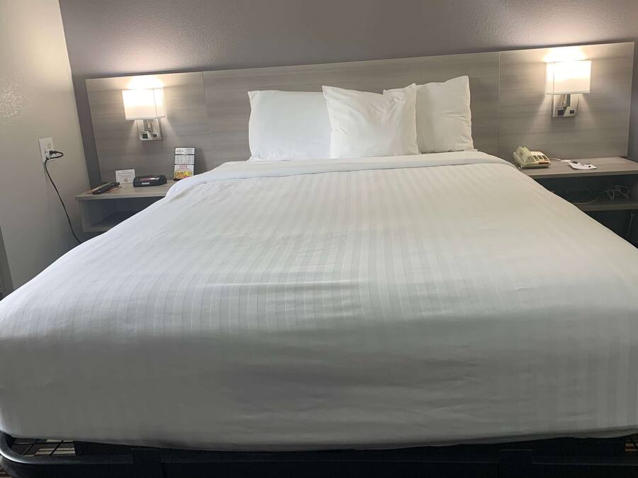 Cama bem arrumada com roupa branca, mesa de cabeceira com abajur, telefone e um aviso na parede.