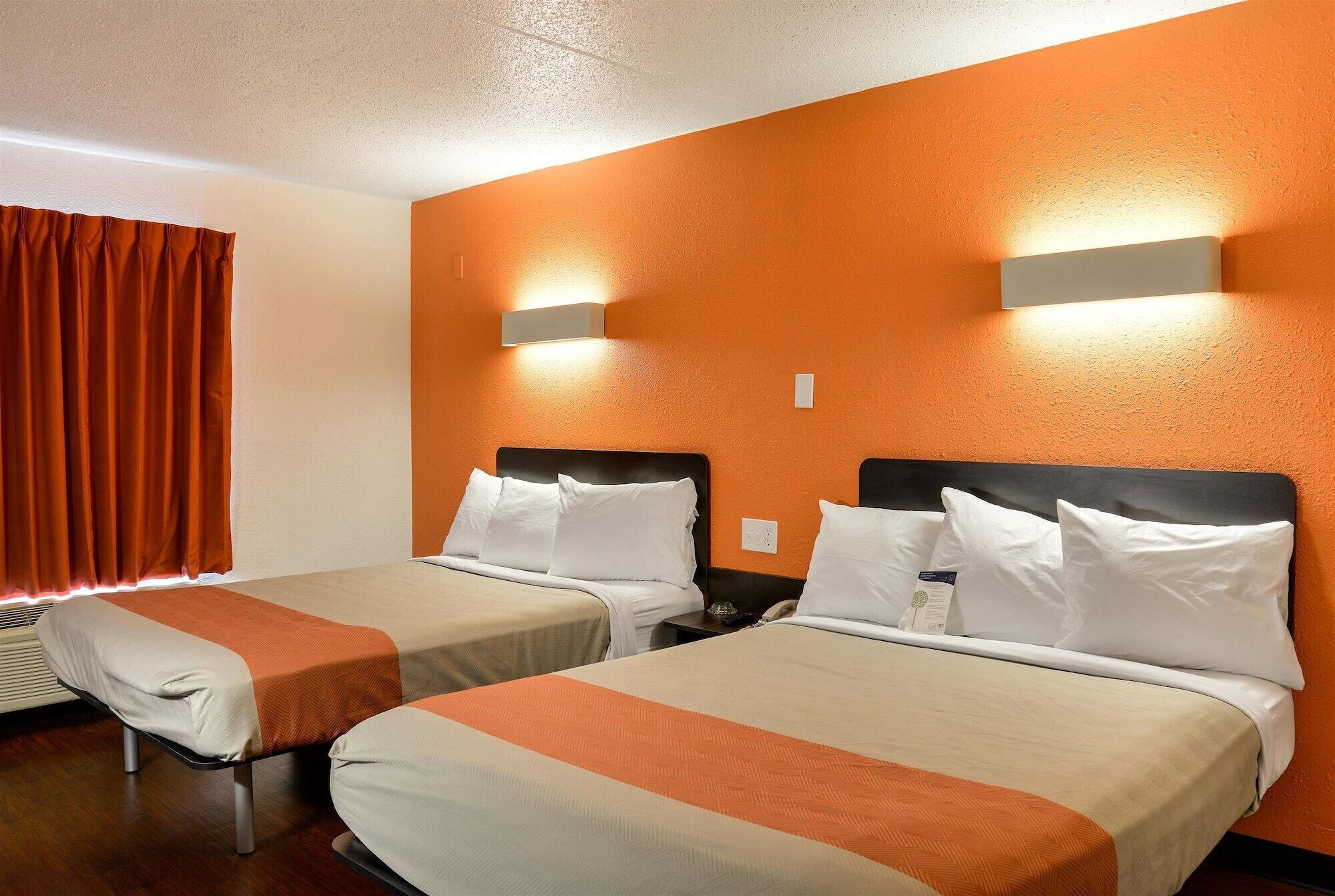A hotel room with two beds, each with white linens and orange accents, and a large orange wall.