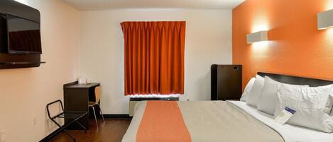 A hotel room with a bed, a desk, a chair, a television, and an orange accent wall.