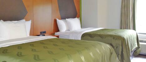 A hotel room with two beds, a green patterned bedspread, a wall with geometric design, and a window with curtains.