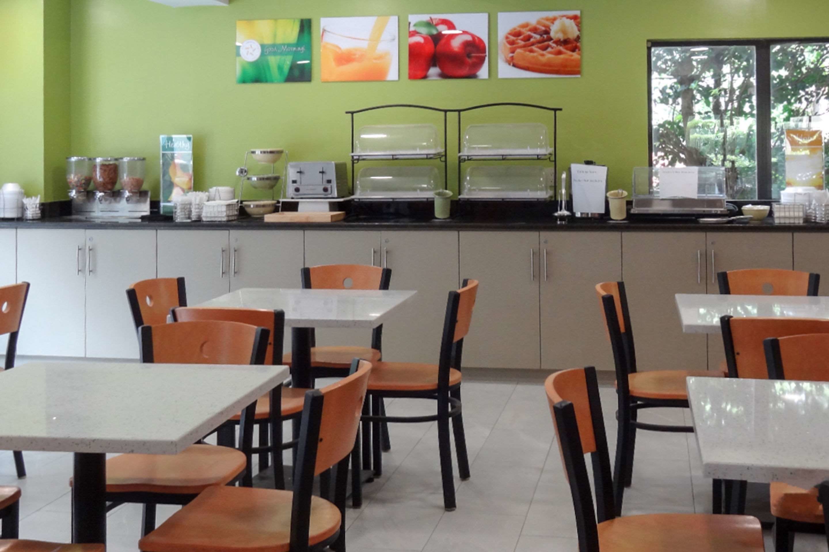 Un restaurant doté de nombreuses tables et chaises, d’un espace buffet avec des récipients contenant de la nourriture, et d’une fenêtre donnant sur des arbres.