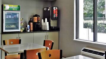 Uma pequena área de refeições com mesa e cadeiras, um refrigerador com bebidas, e uma janela com vista para árvores.