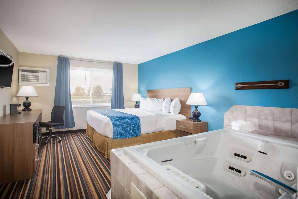 Habitación de hotel con cama, escritorio, silla, lámpara de escritorio, mesita de noche con lámpara, ventana con cortinas, bañera y vistas al exterior.