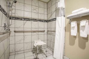 A bathroom with white marble tiles, a shower with a handheld showerhead, a grab bar, a white portable toilet, and a towel rack with folded towels. - Travelodge by Wyndham Wahpeton Event Center (Wahpeton)