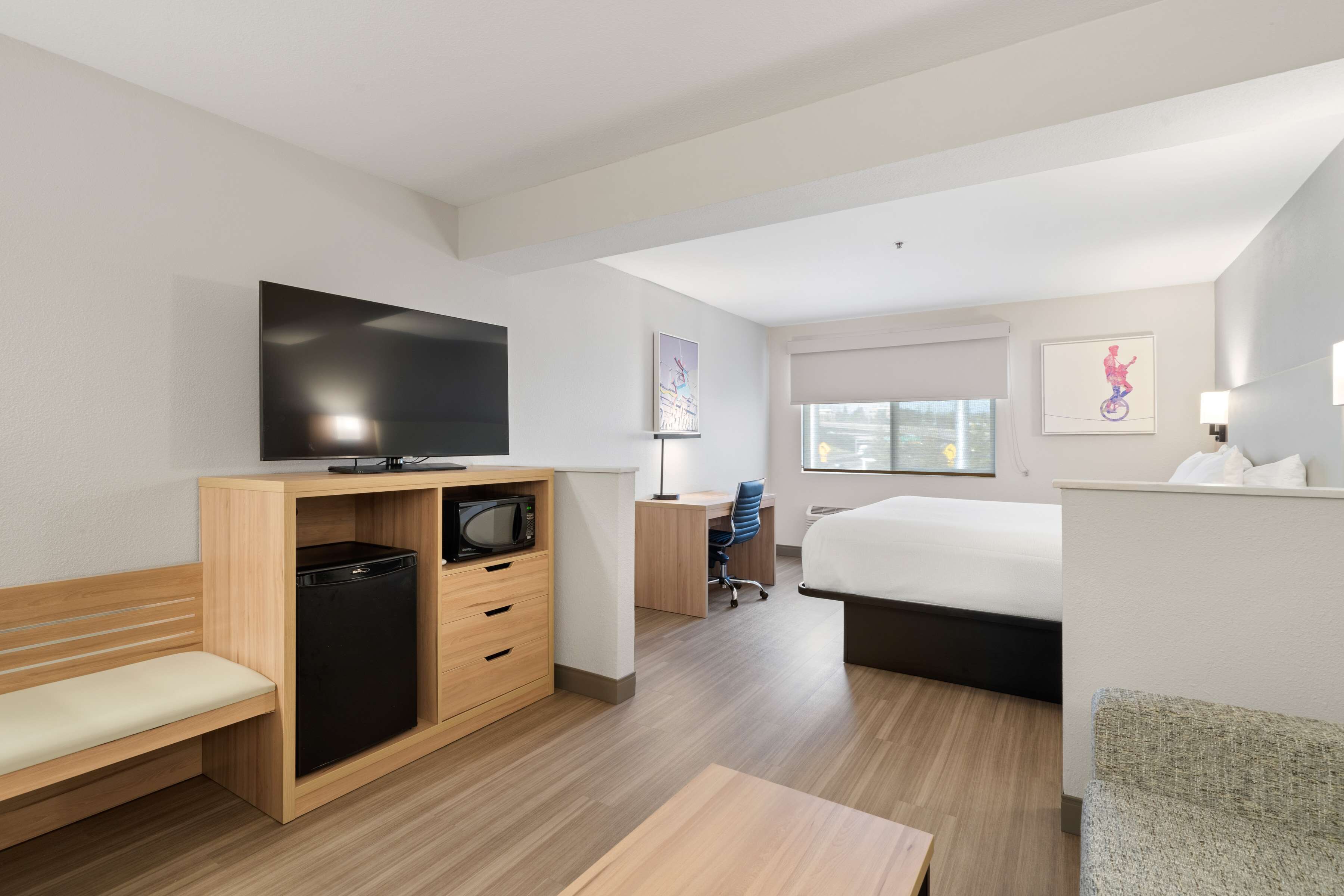 A modern hotel room with a bed, a desk with a computer, a television, and a bench.