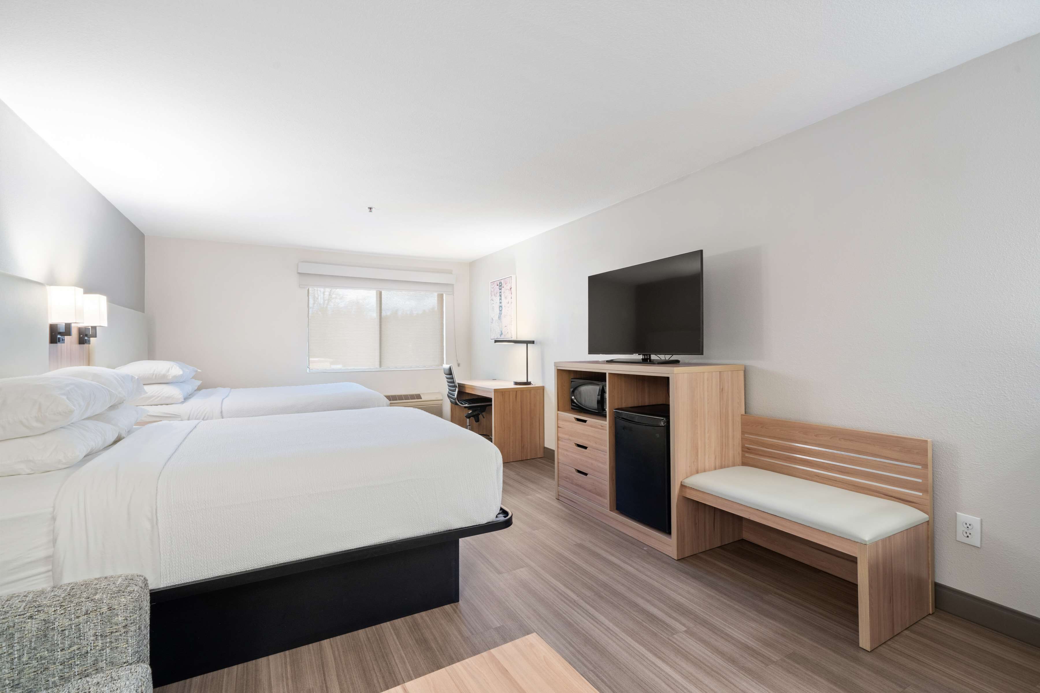 A modern hotel room with a large bed, a desk, a television, and a bench.