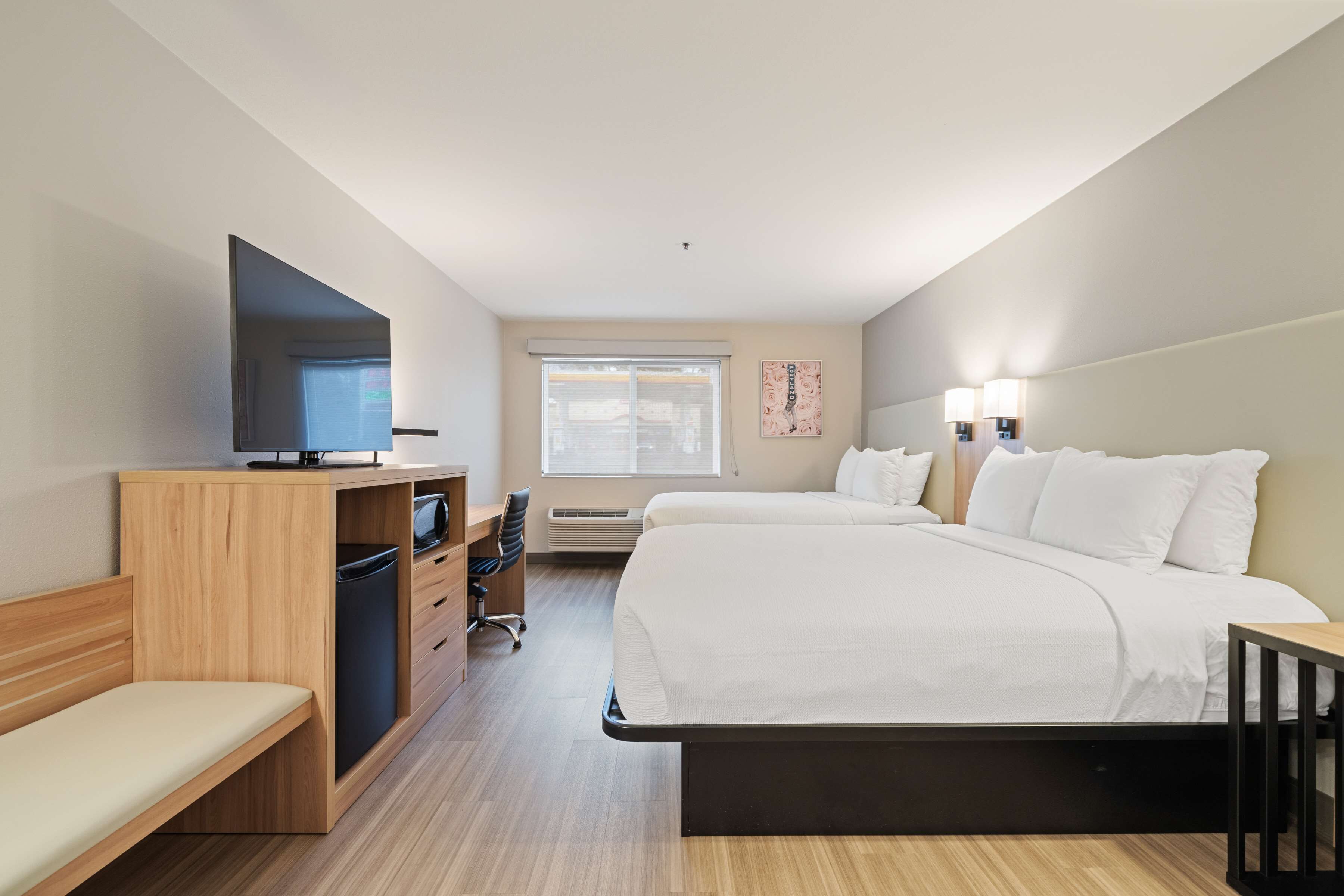 Habitación de hotel con cama, escritorio con computadora, televisor, ventana y una obra de arte colgada en la pared.