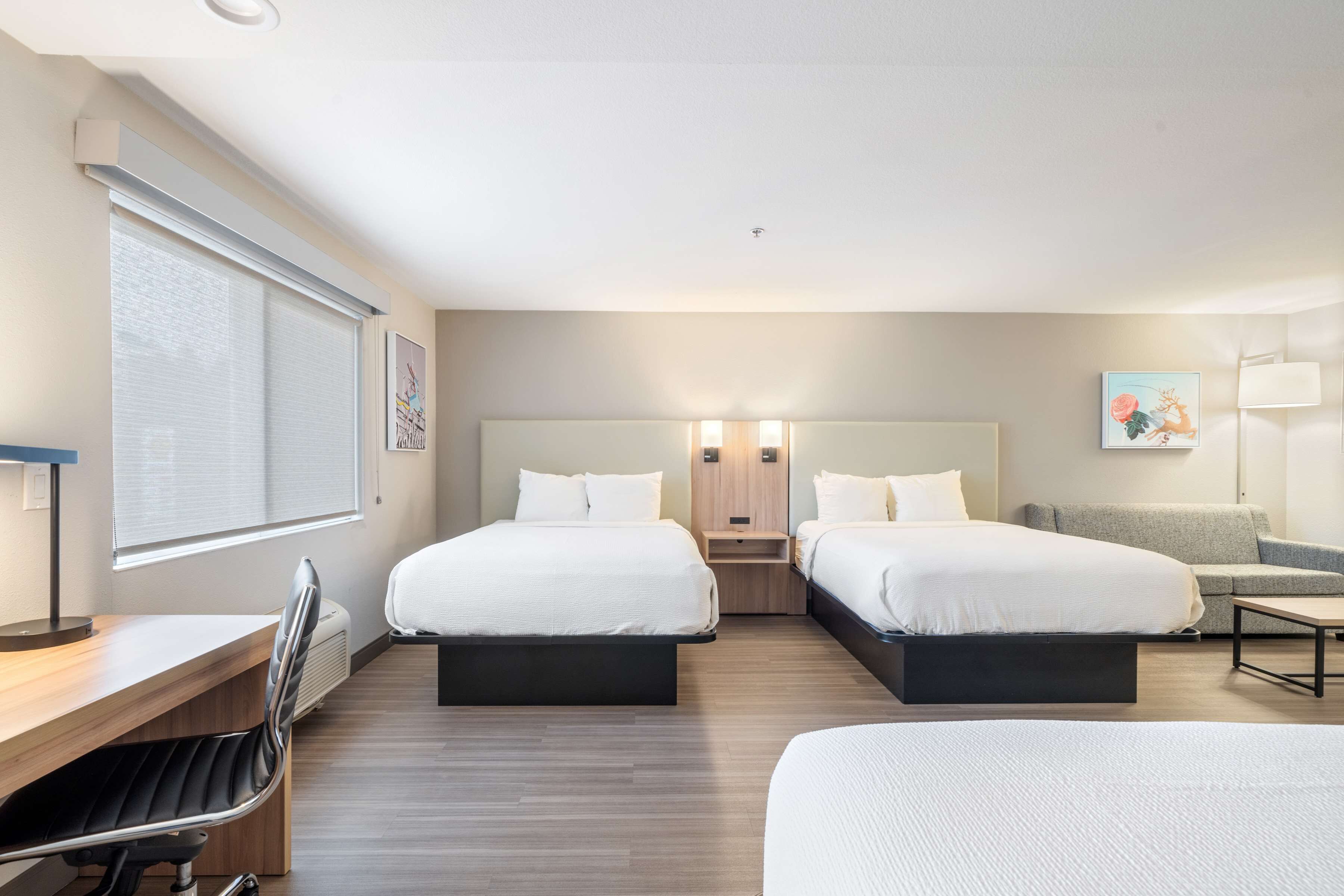 A hotel room with two beds, a desk, a chair, a sofa, and a window with blinds.