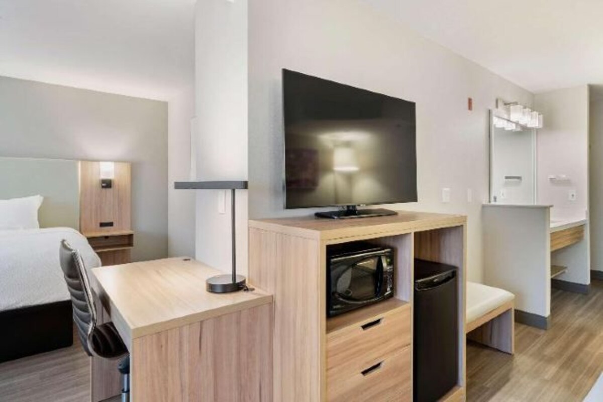 A hotel room with a bed, a desk with a TV, a microwave, and a bathroom with a mirror.