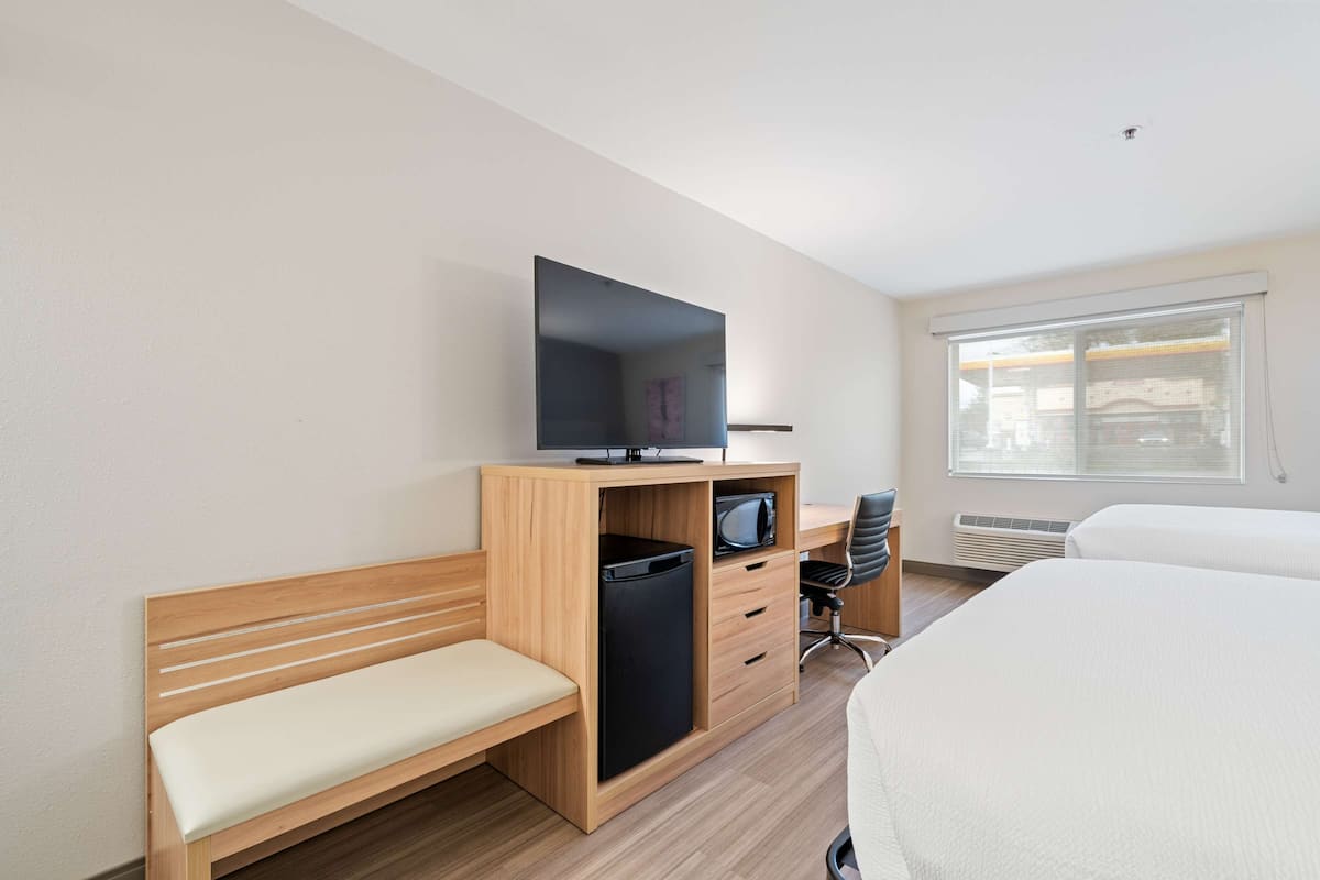 A hotel room with a wooden desk, a television, a small trash bin, a chair, a bed, and a window with blinds.