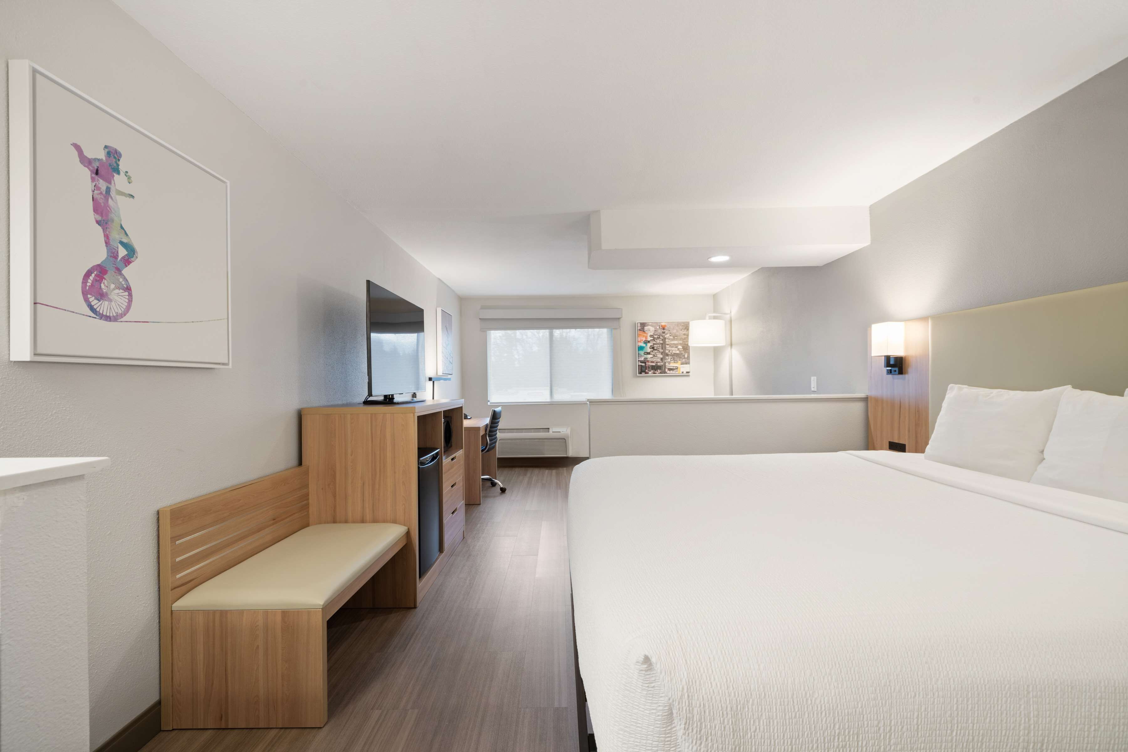 A hotel room with a large bed, a wooden bench, a desk with a chair, a TV, and a window with blinds.