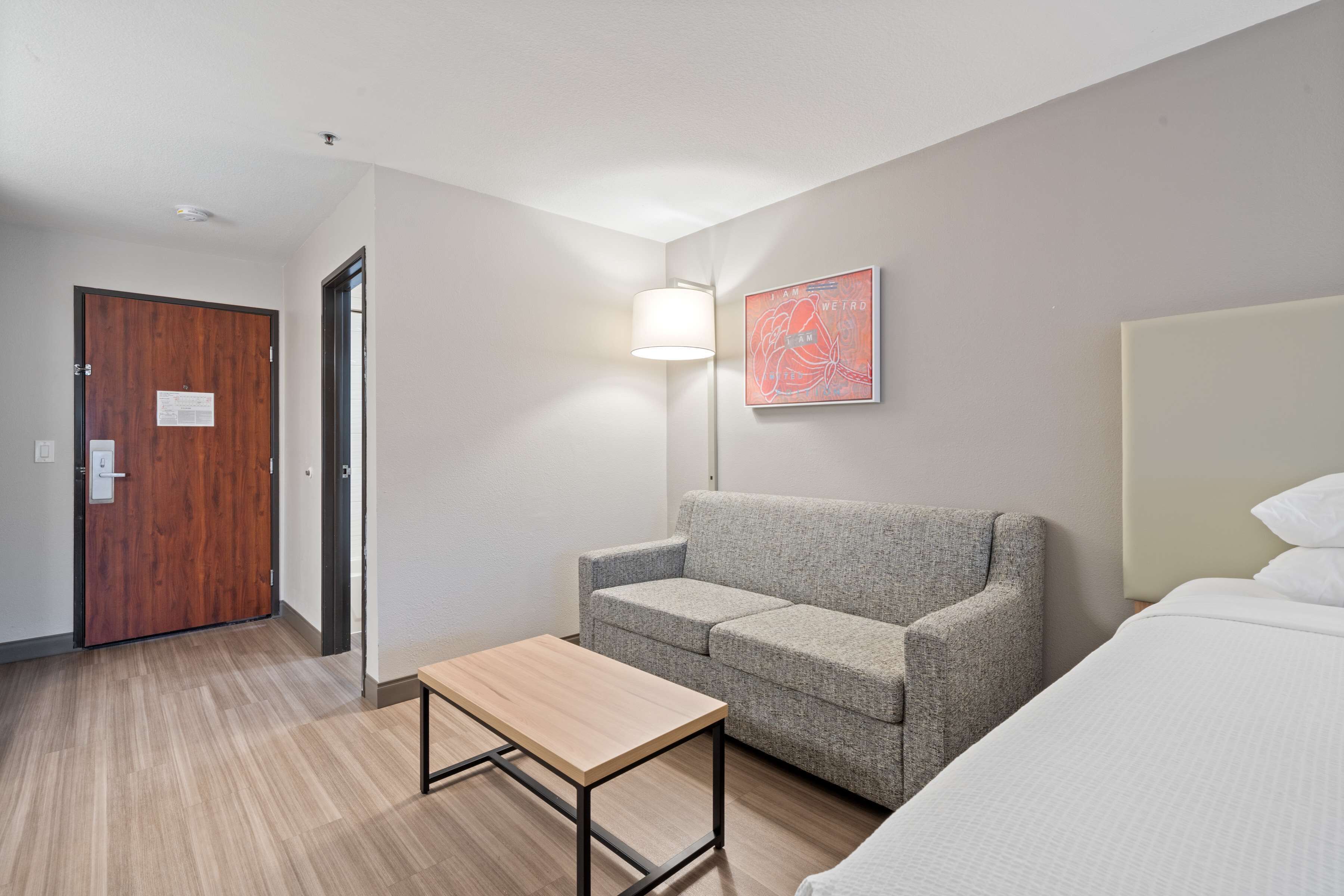 A hotel room with a wooden door, a grey sofa, a small wooden table, a white bed, and a wall-mounted lamp.
