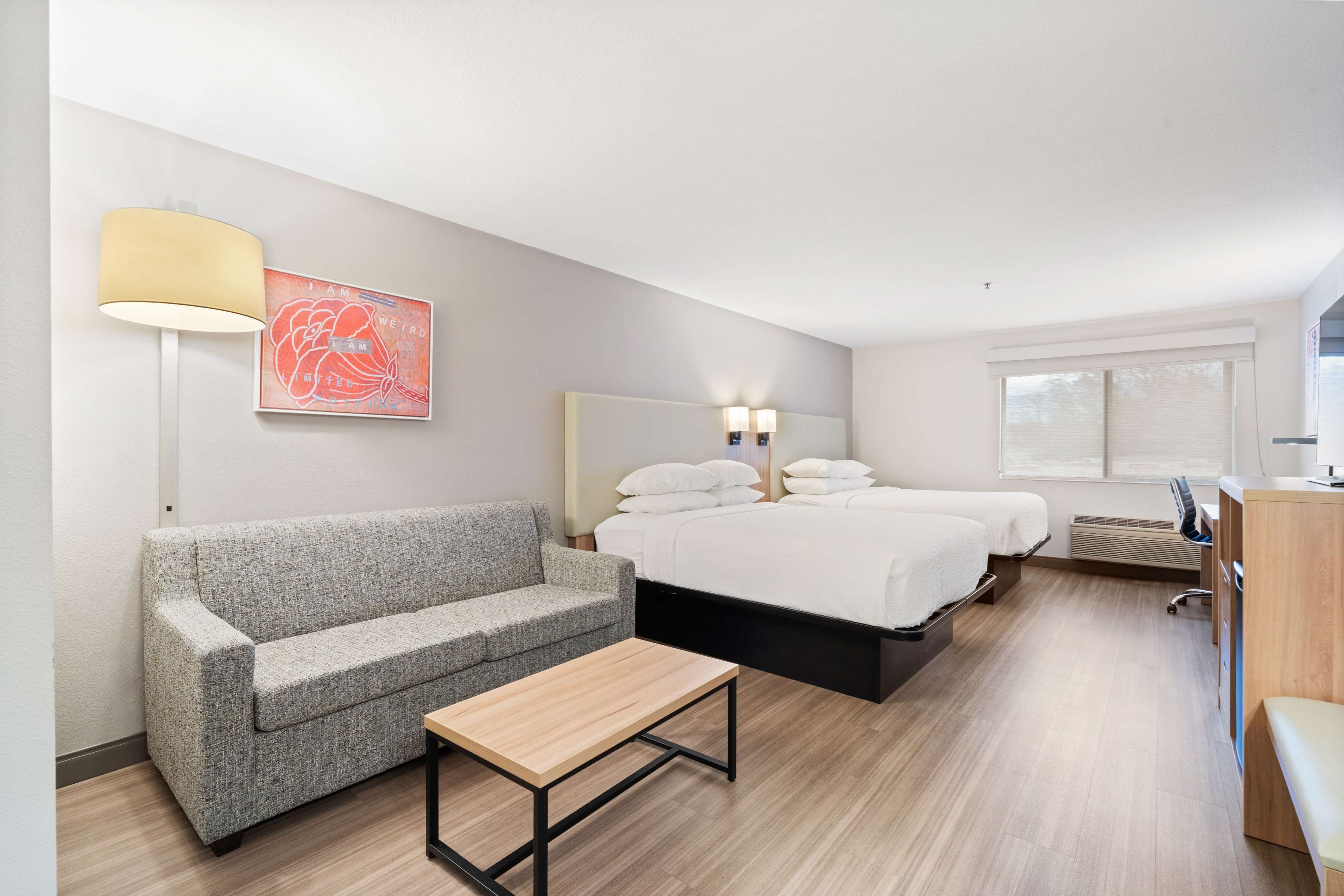 A modern hotel room with a grey sofa, a wooden table, a bed with white linens, a wall-mounted lamp, and a framed artwork.