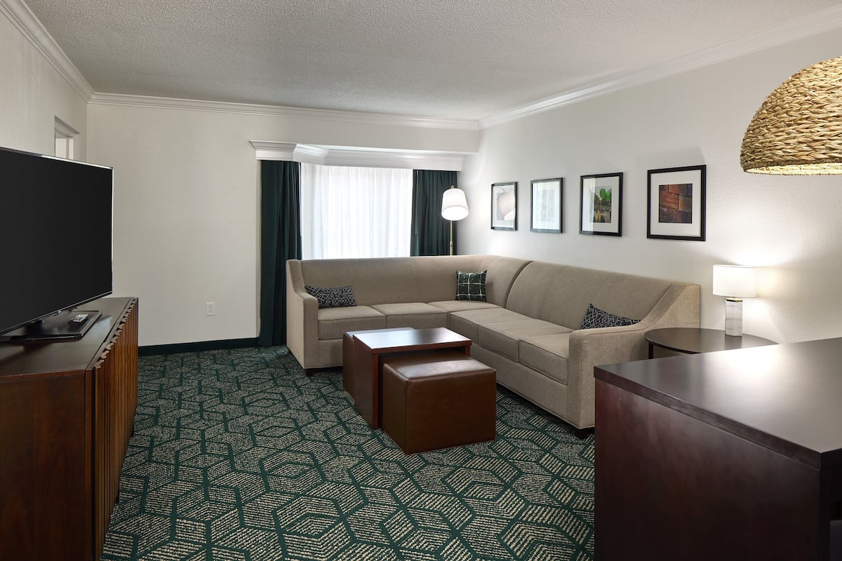A hotel room with a beige sofa, a wooden coffee table, a TV on a wooden stand, and a patterned carpet.