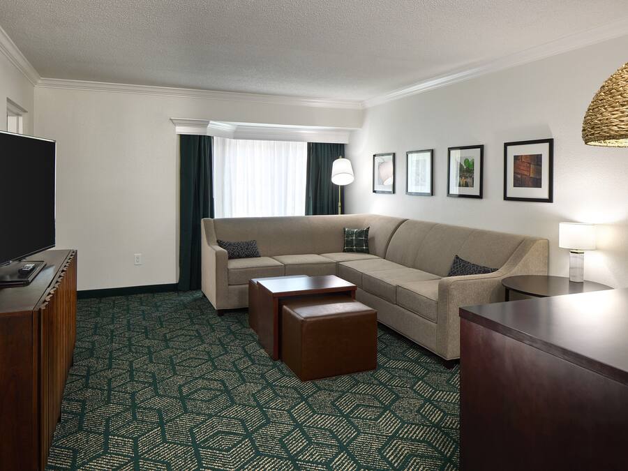 Une chambre d’hôtel avec un canapé beige, une table basse en bois, un téléviseur posé sur un meuble en bois et une moquette à motifs.