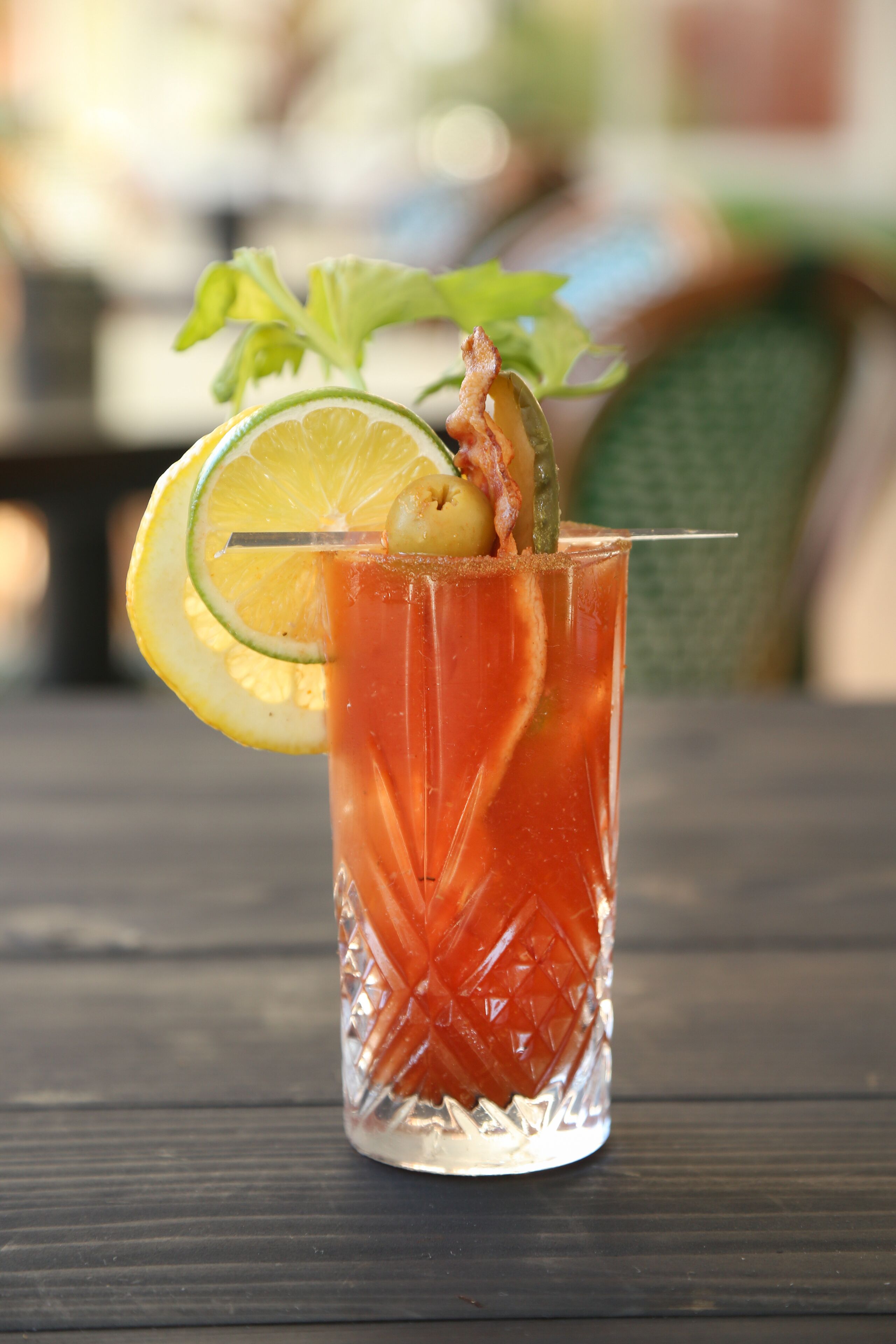 Un verre de Bloody Mary, garni d’une tranche de citron vert et accompagné de quelques tranches de bacon, sur une table en bois.