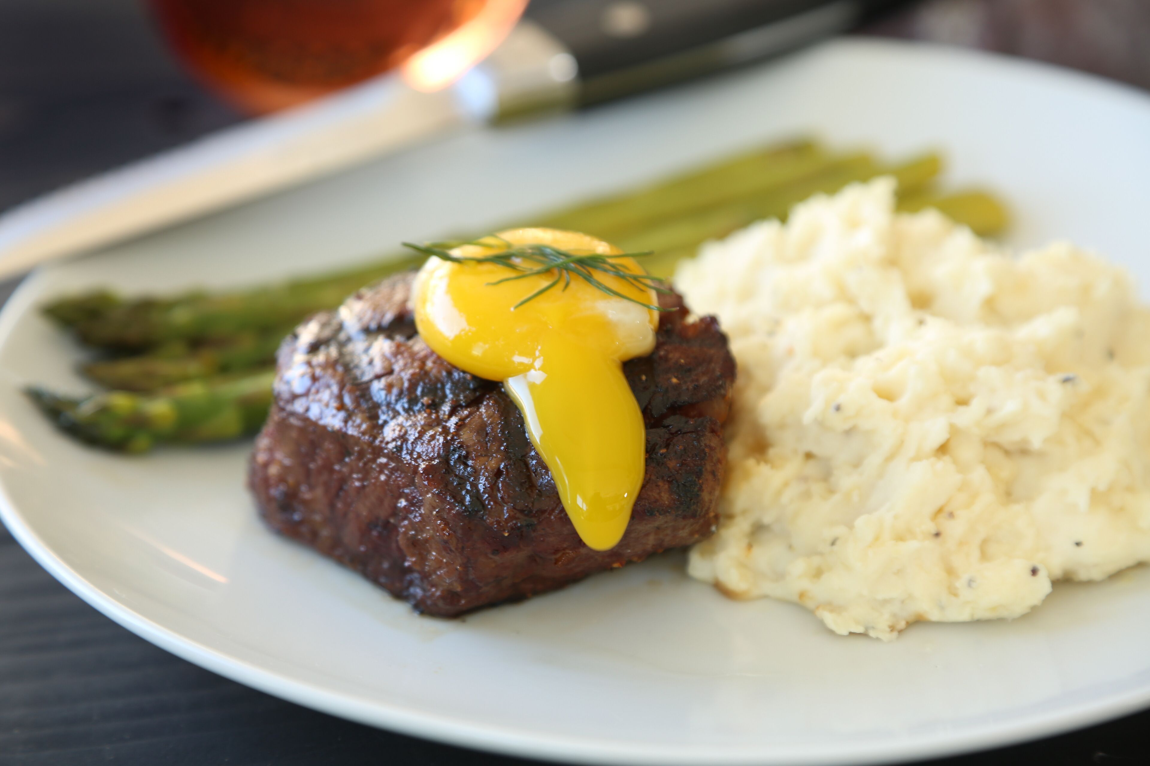 A plate with a steak topped with a yellow sauce, asparagus, and mashed potatoes.
