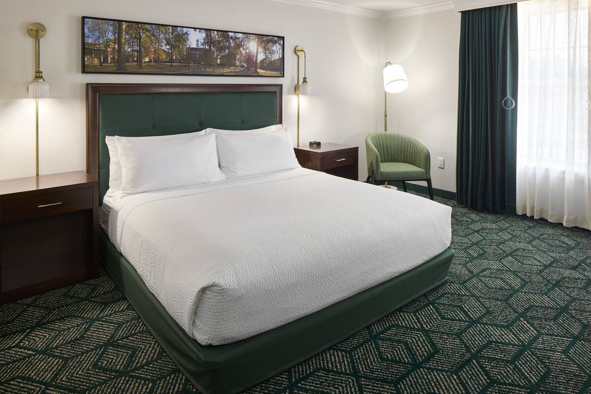 Habitació d'hotel amb un llit gran, un panell de capçalera verd, una taula de nit, una cadira verda, una làmpada i una finestra amb cortines.
