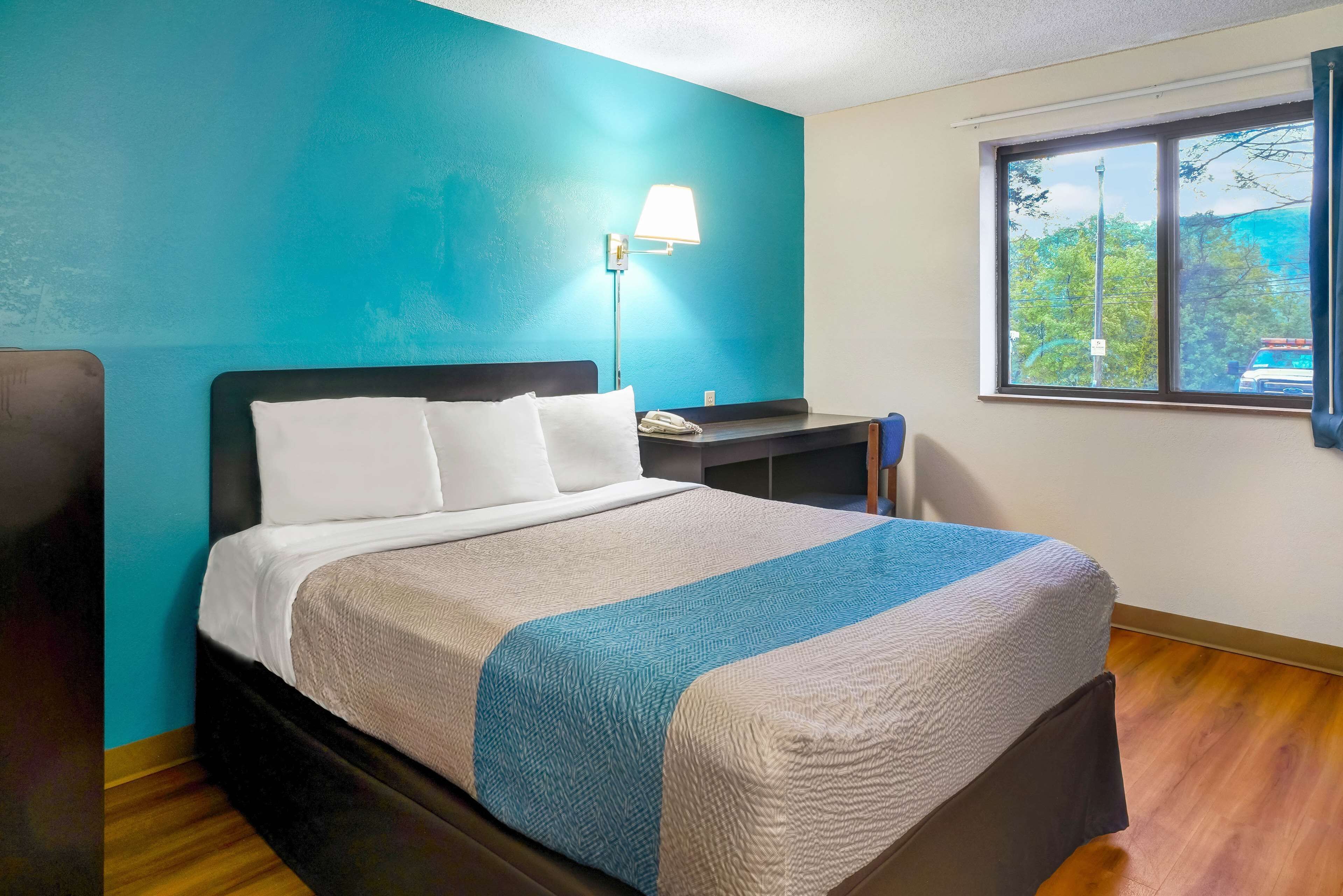 A neatly made bed with white and blue bedding, a bedside lamp, and a window with a view of trees.