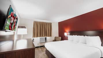 A hotel room with a bed, a desk, a television, a window with curtains, and a wall-mounted air conditioning unit.