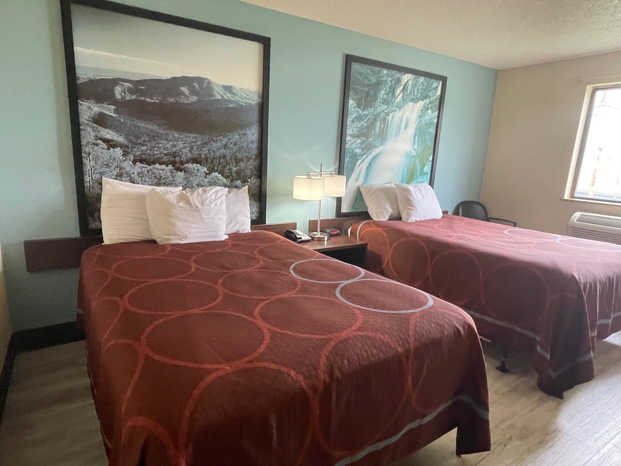 Habitación de hotel con dos camas, cada una con una colcha estampada roja, una mesita de noche con lámpara, y dos cuadros enmarcados en la pared.
