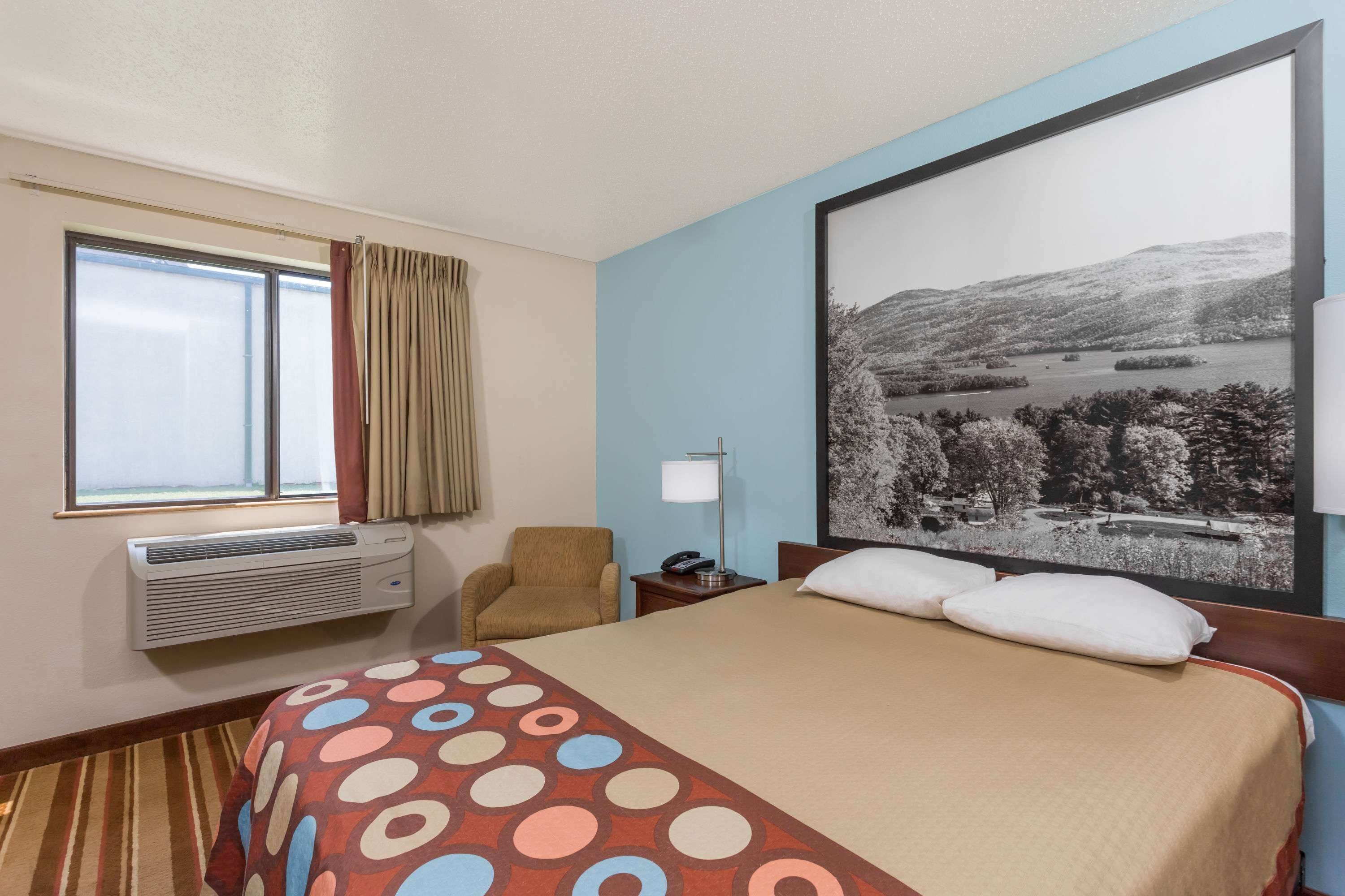 A hotel room with a bed, a chair, a window with curtains, an air conditioner, a nightstand with a lamp, and a large framed picture on the wall.