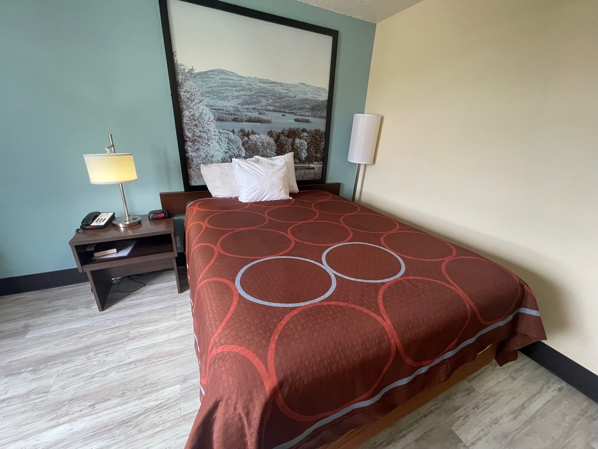 Een hotelkamer met een bed, nachtkastje, lamp, telefoon en een schilderij aan de muur.