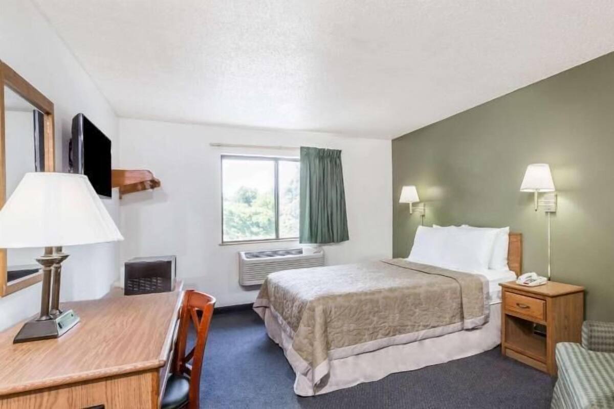 Habitación de hotel con cama, mesita de noche, lámpara, silla, escritorio y ventana con cortinas.