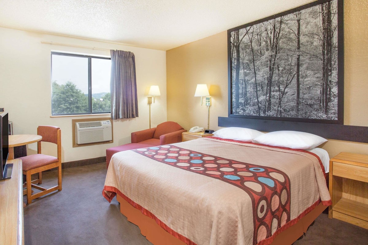 A hotel room with a bed, a desk, a chair, a window with curtains, an air conditioner, two bedside lamps, and a large framed picture on the wall.