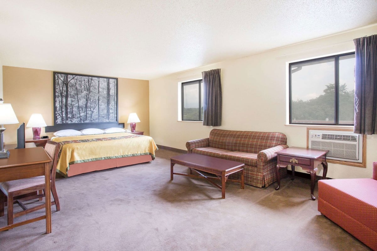A hotel room with a bed, a sofa, a table, and a chair. There is a window with curtains, a wall-mounted air conditioner, and a painting on the wall.