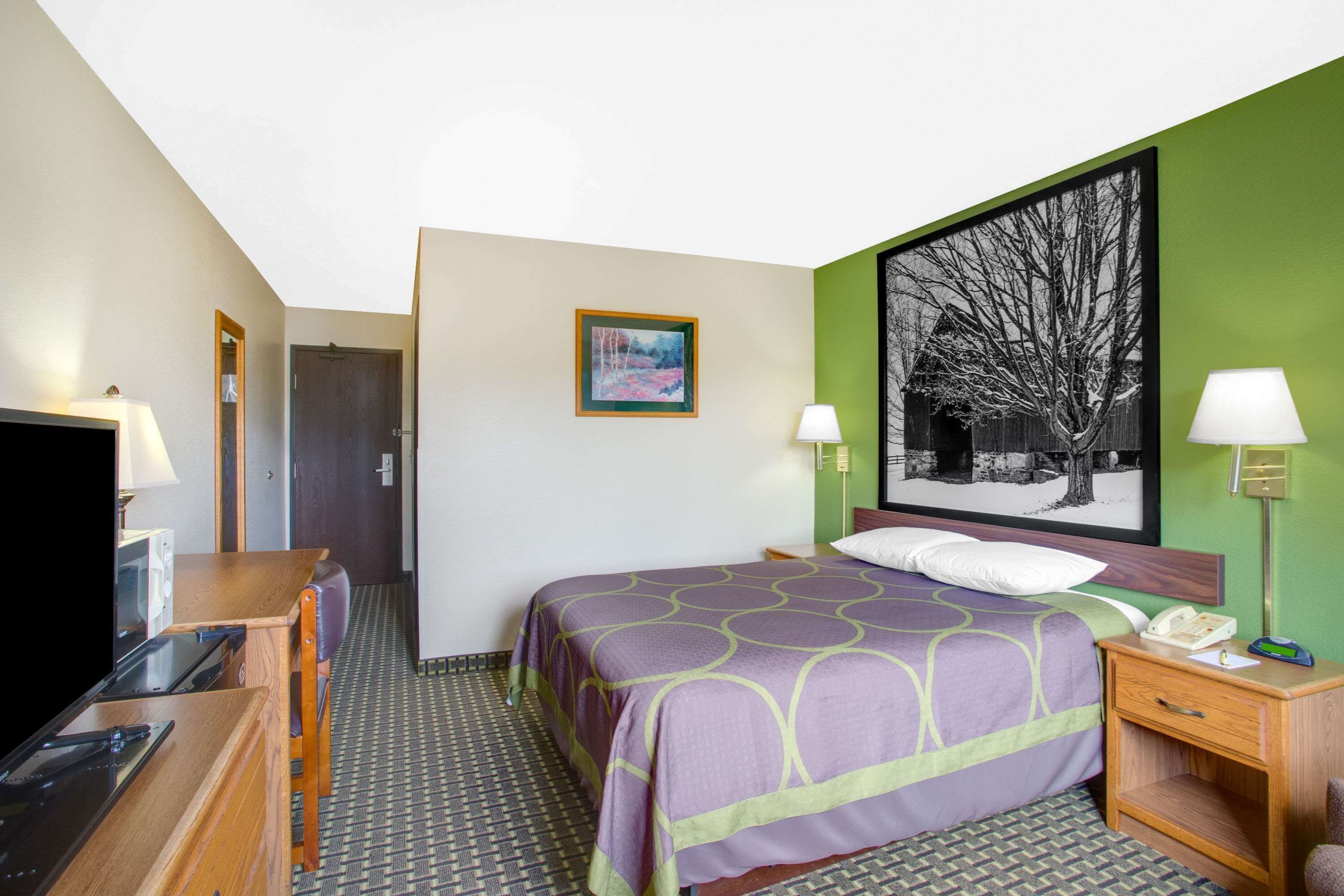 A hotel room with a bed, bedside table, lamp, and a large framed picture on the wall.