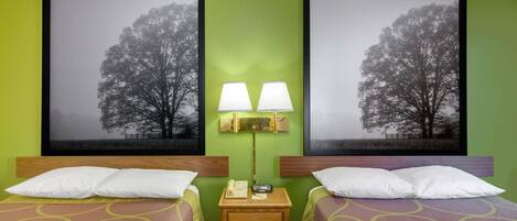 A hotel room with two beds, a nightstand with a lamp, and two framed pictures of trees on the wall.