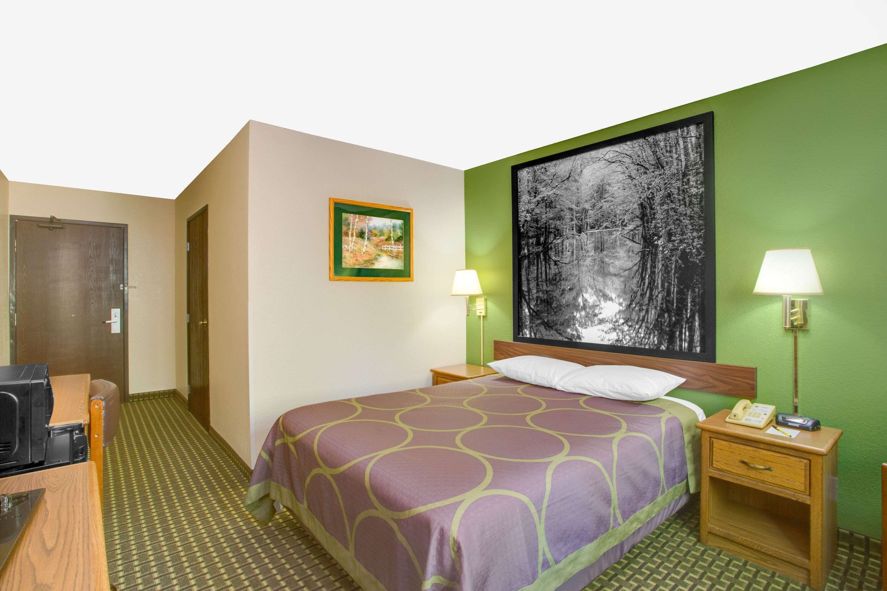 Una camera d'albergo con un letto, due lampade ai piedi del letto, un comodino con il telefono, un quadro appeso alla parete e una porta.