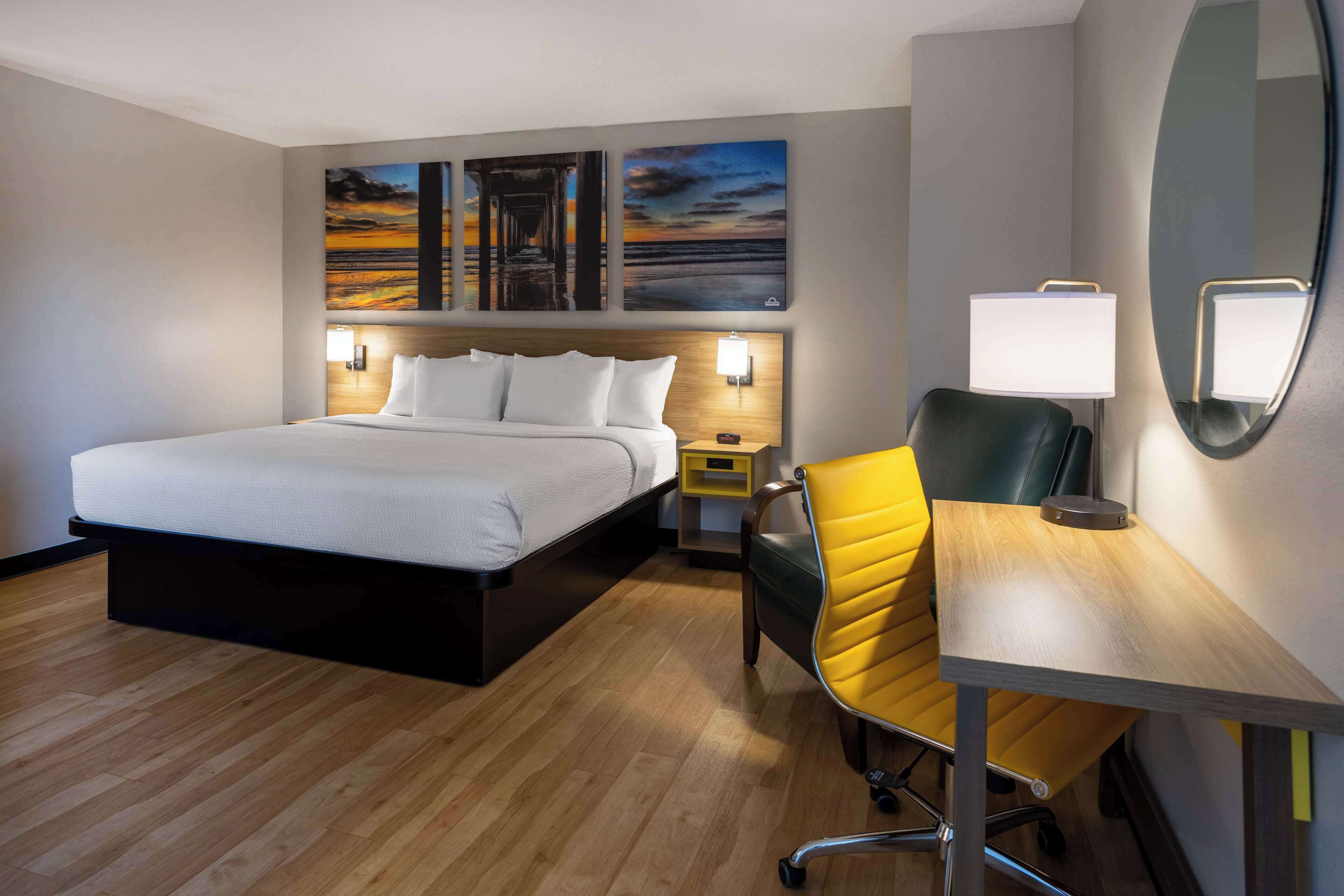 A hotel room with a bed, a desk, a chair, and artwork on the wall.