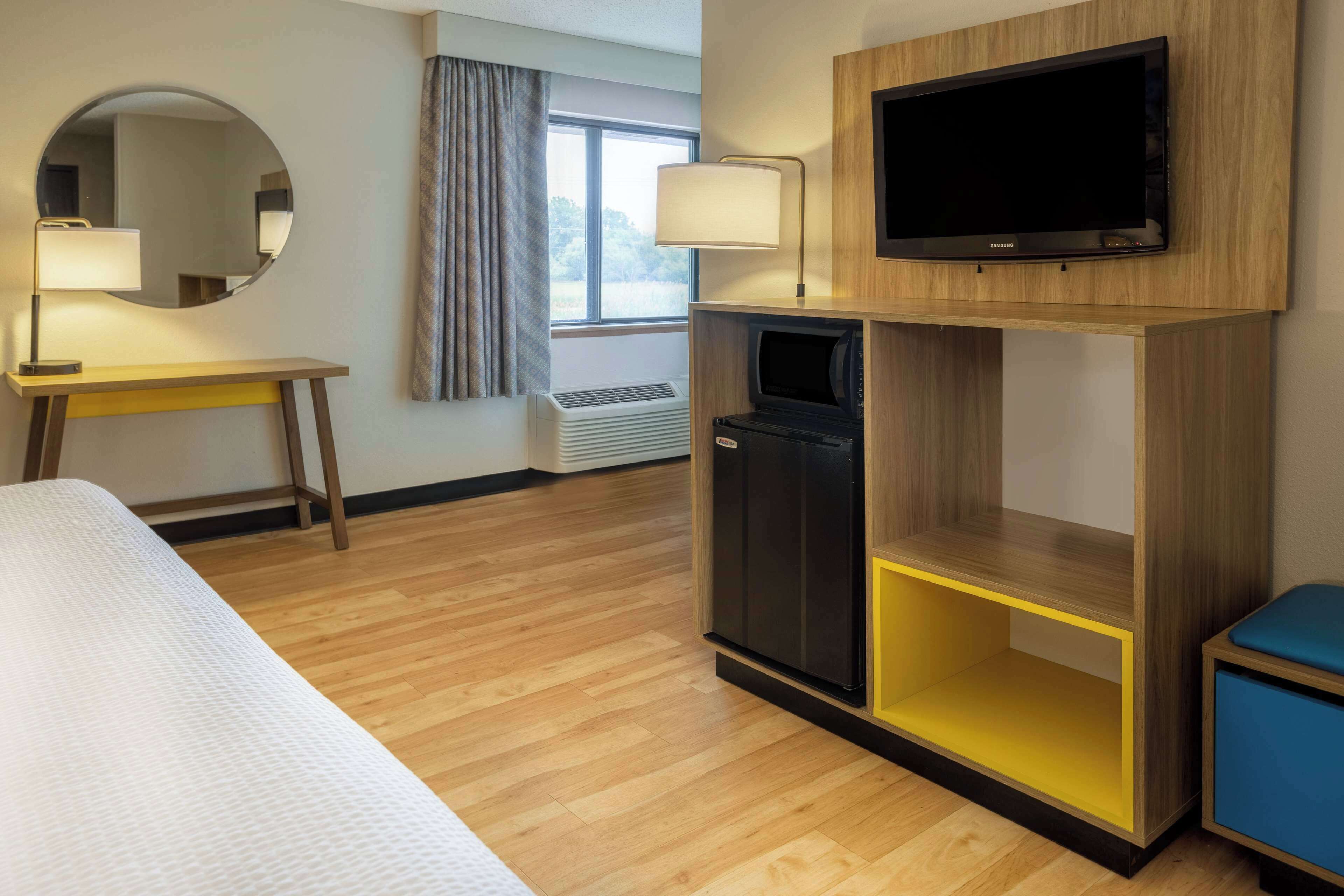 A hotel room with a bed, a desk with a lamp, a television on a wooden stand, a small cabinet, and a window with curtains.