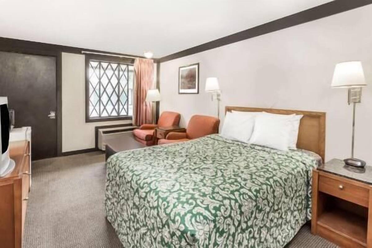 A hotel room with a large bed, a desk, a chair, a window with curtains, and a wall-mounted lamp.
