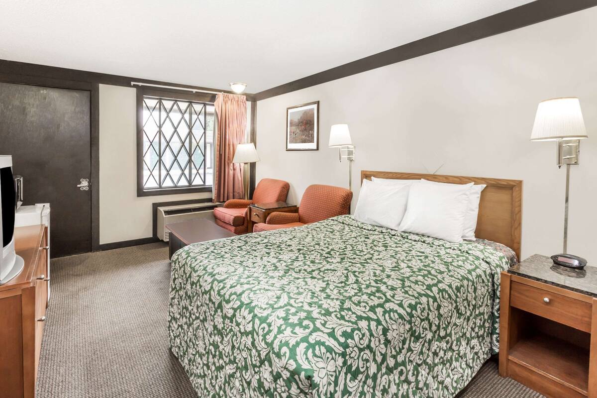 A hotel room with a bed, two chairs, a desk, a lamp, and a television.