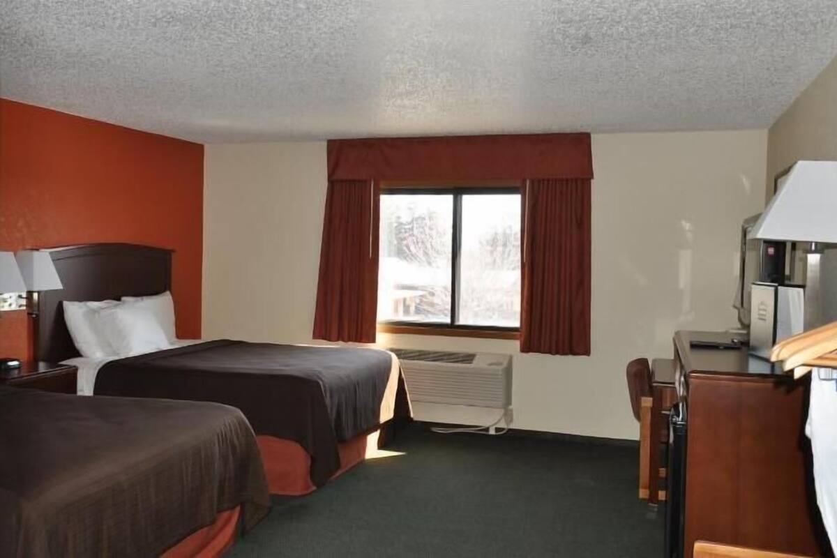 A hotel room with two beds, a desk, a chair, a lamp, and a window with curtains.