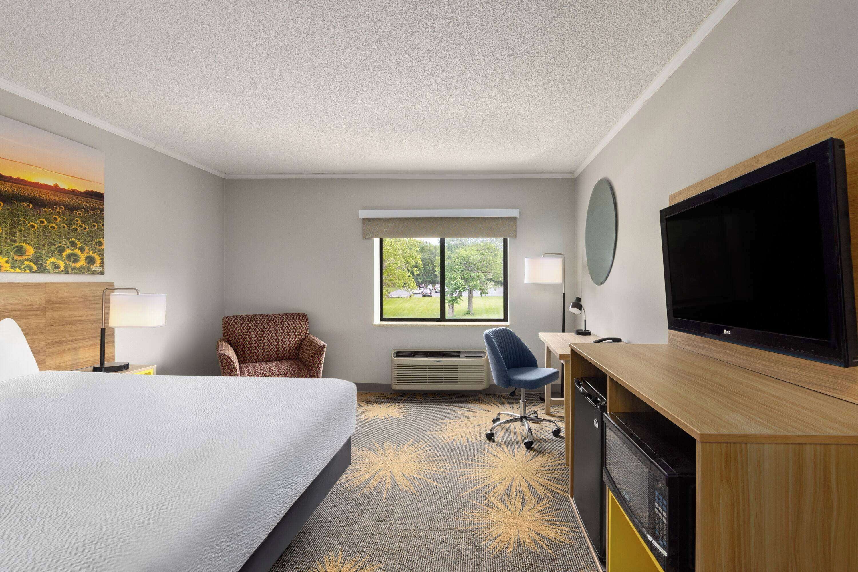 A hotel room with a bed, a desk with a television, a chair, a window with a view of greenery, and a wall-mounted mirror.