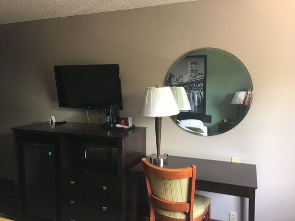 Ein Hotelzimmer mit Flachbildfernseher, Kaffeemaschine, Lampe, Stuhl, Schreibtisch und rundem Spiegel.