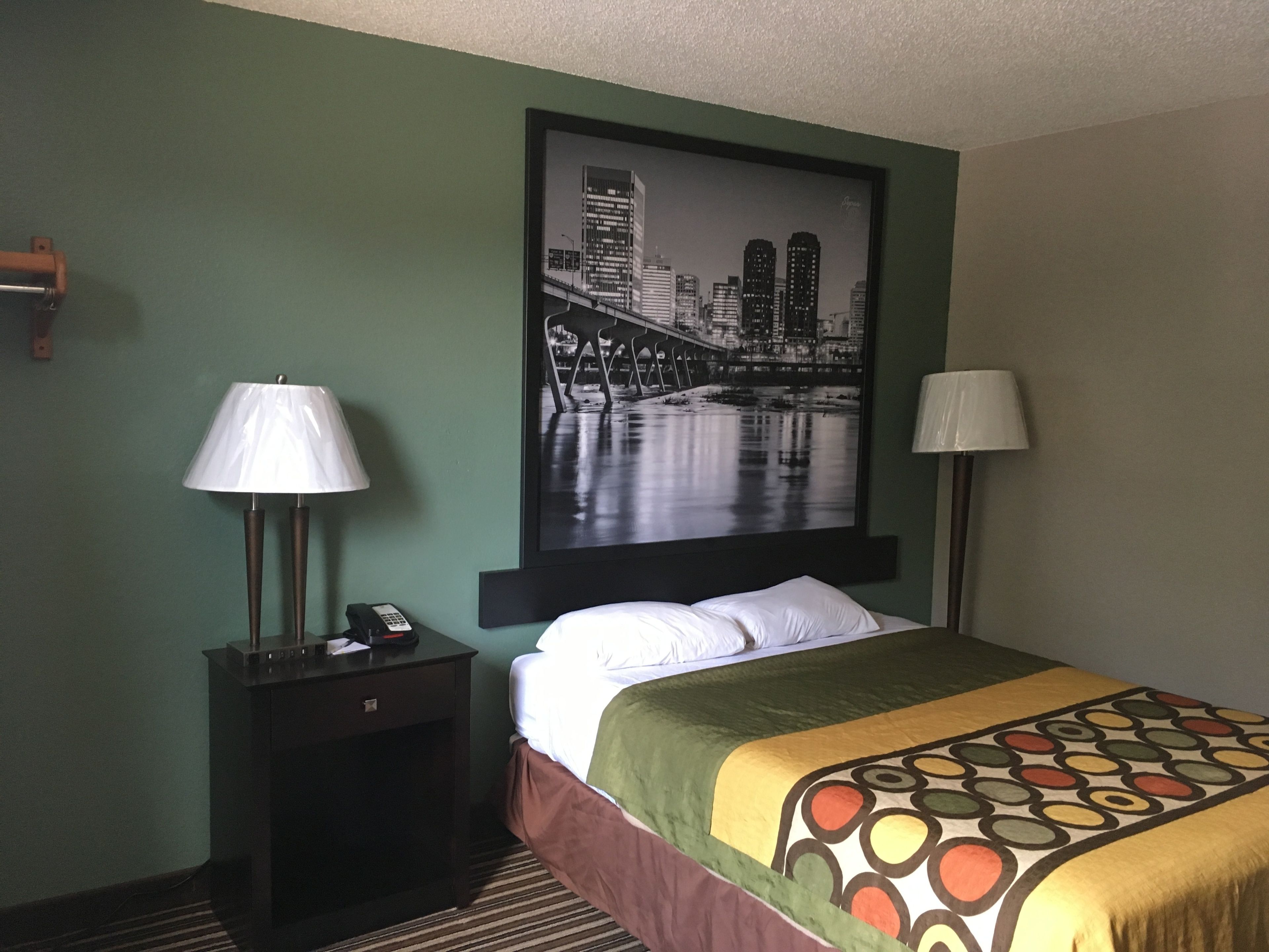 Una camera d'albergo con un letto, due lampade sui comodini, un comodino con un telefono e un quadro con una veduta cittadina appeso al muro.