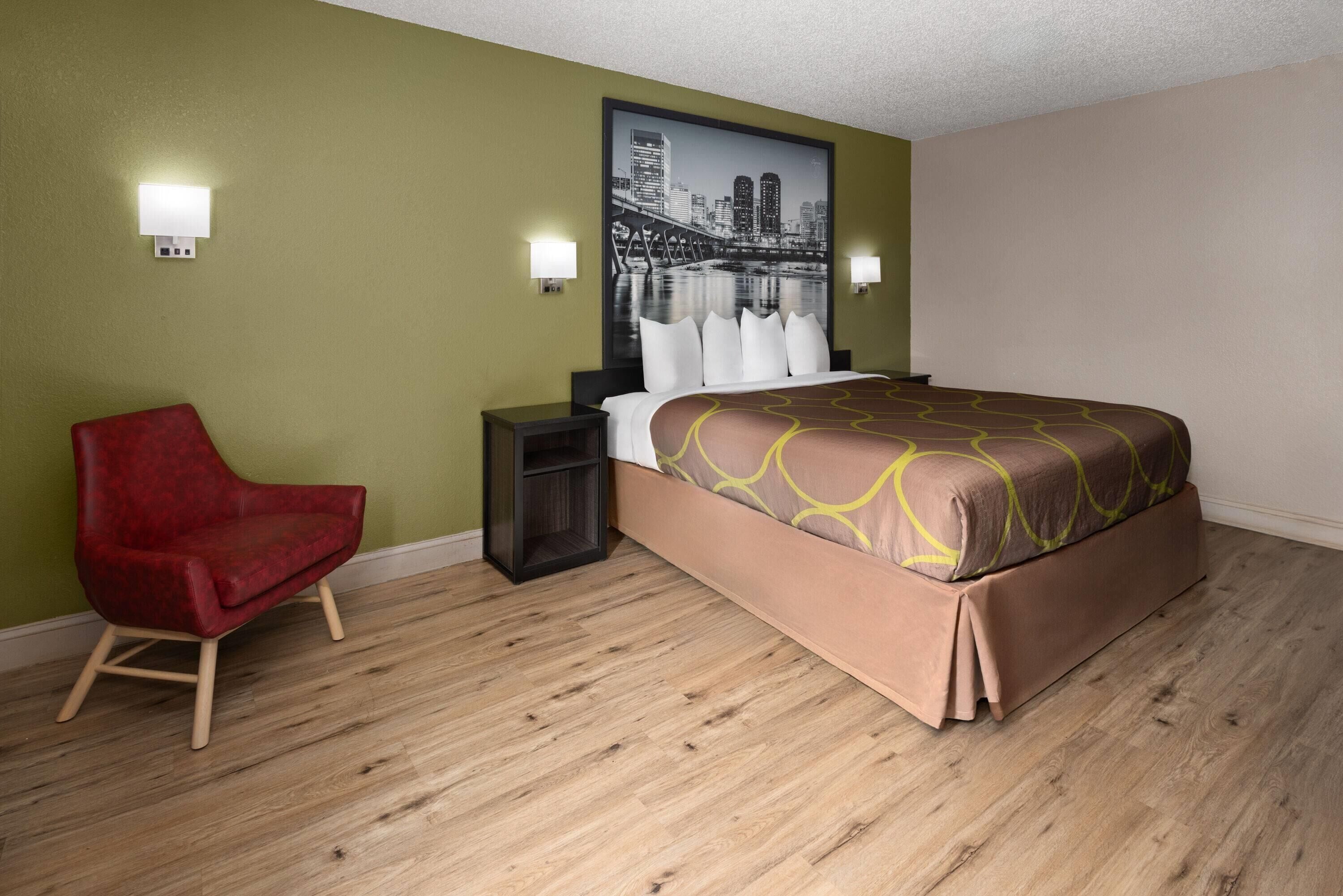 A hotel room with a bed, a red chair, a nightstand, and a wall-mounted light fixture.