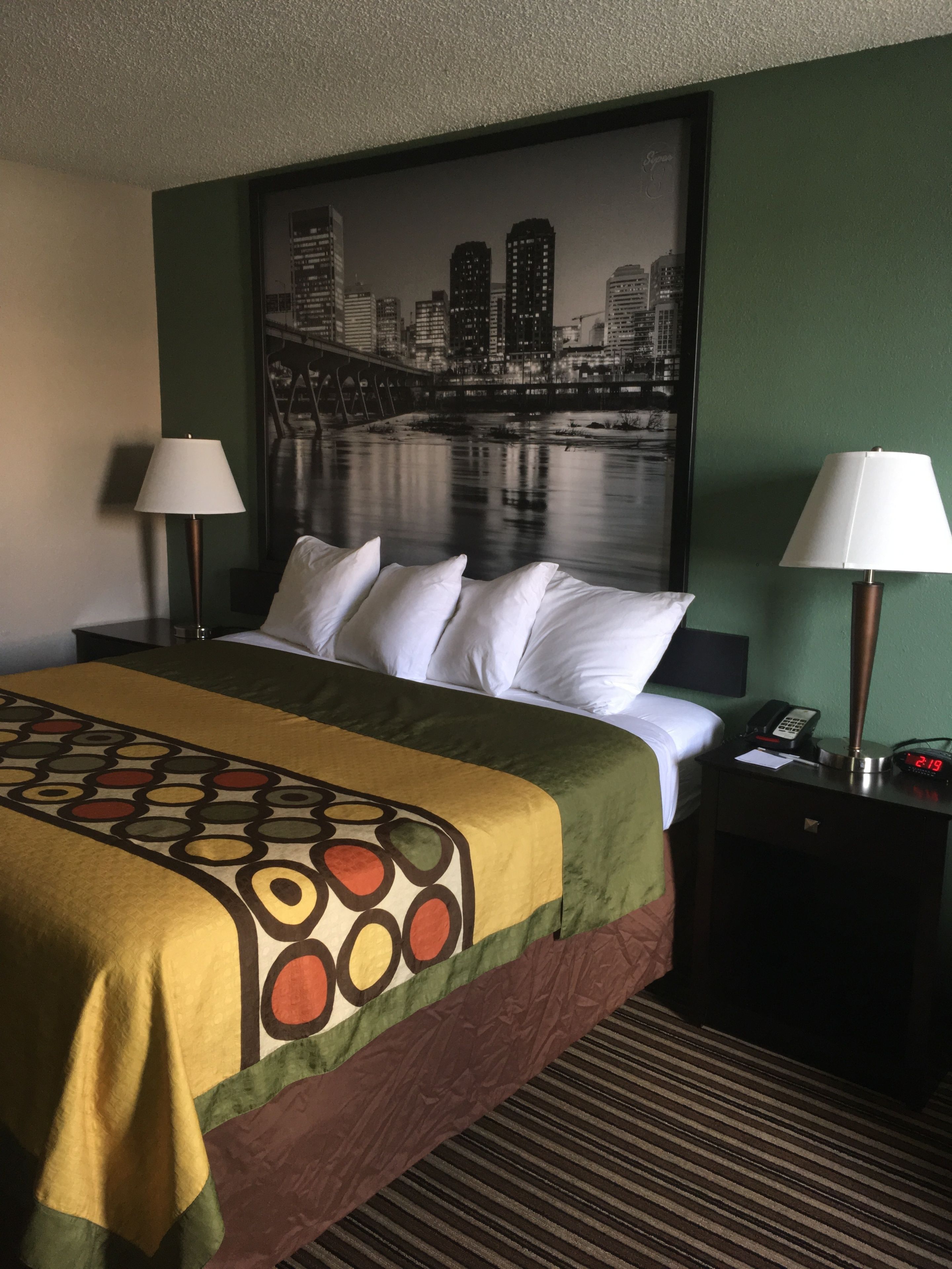 A hotel room with a bed, two bedside tables, a cityscape painting, and a clock.