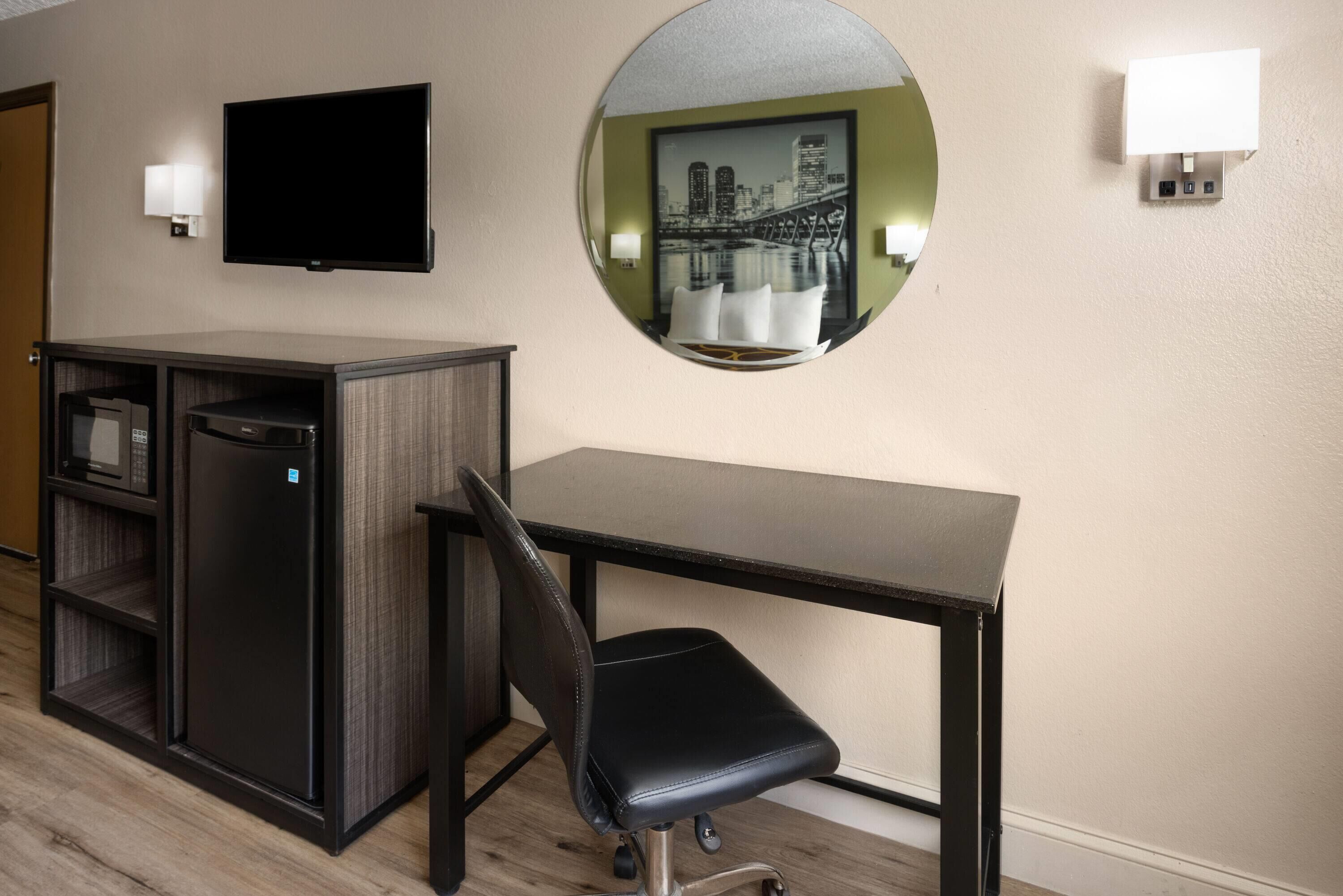 A hotel room with a desk, chair, television, minibar, and a round mirror on the wall.