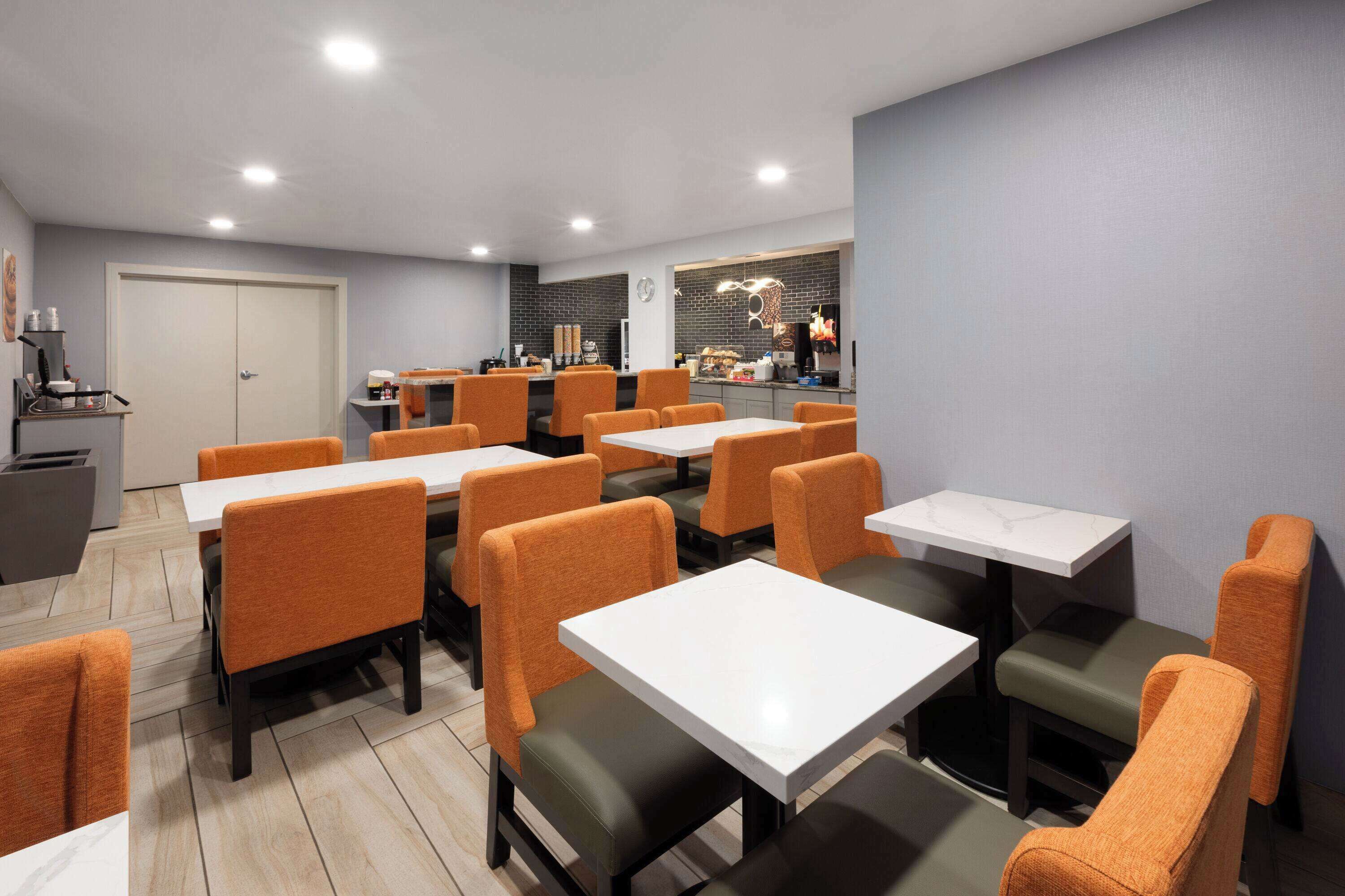 A modern restaurant with multiple tables and orange chairs, featuring a brick wall and a visible kitchen area.