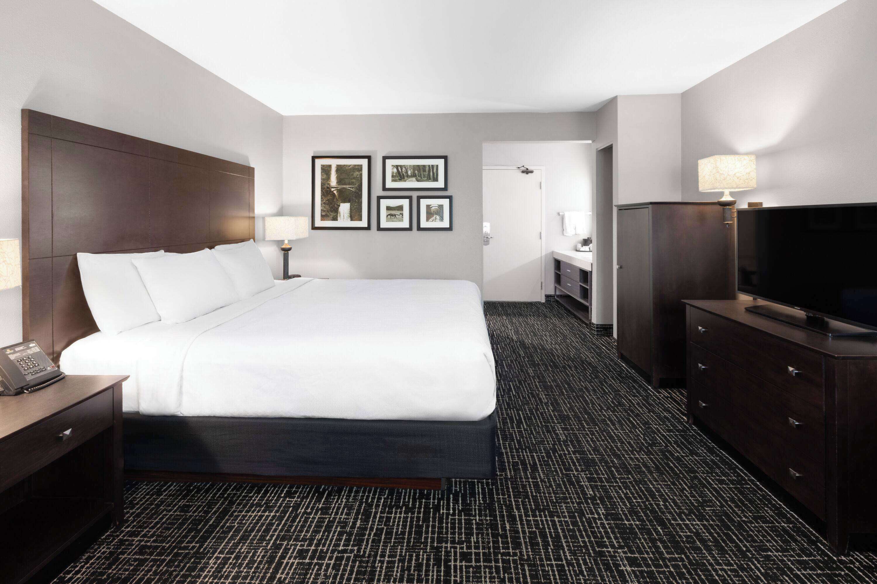 A hotel room with a large bed, a desk with a telephone, a dresser with a television, and framed pictures on the wall.