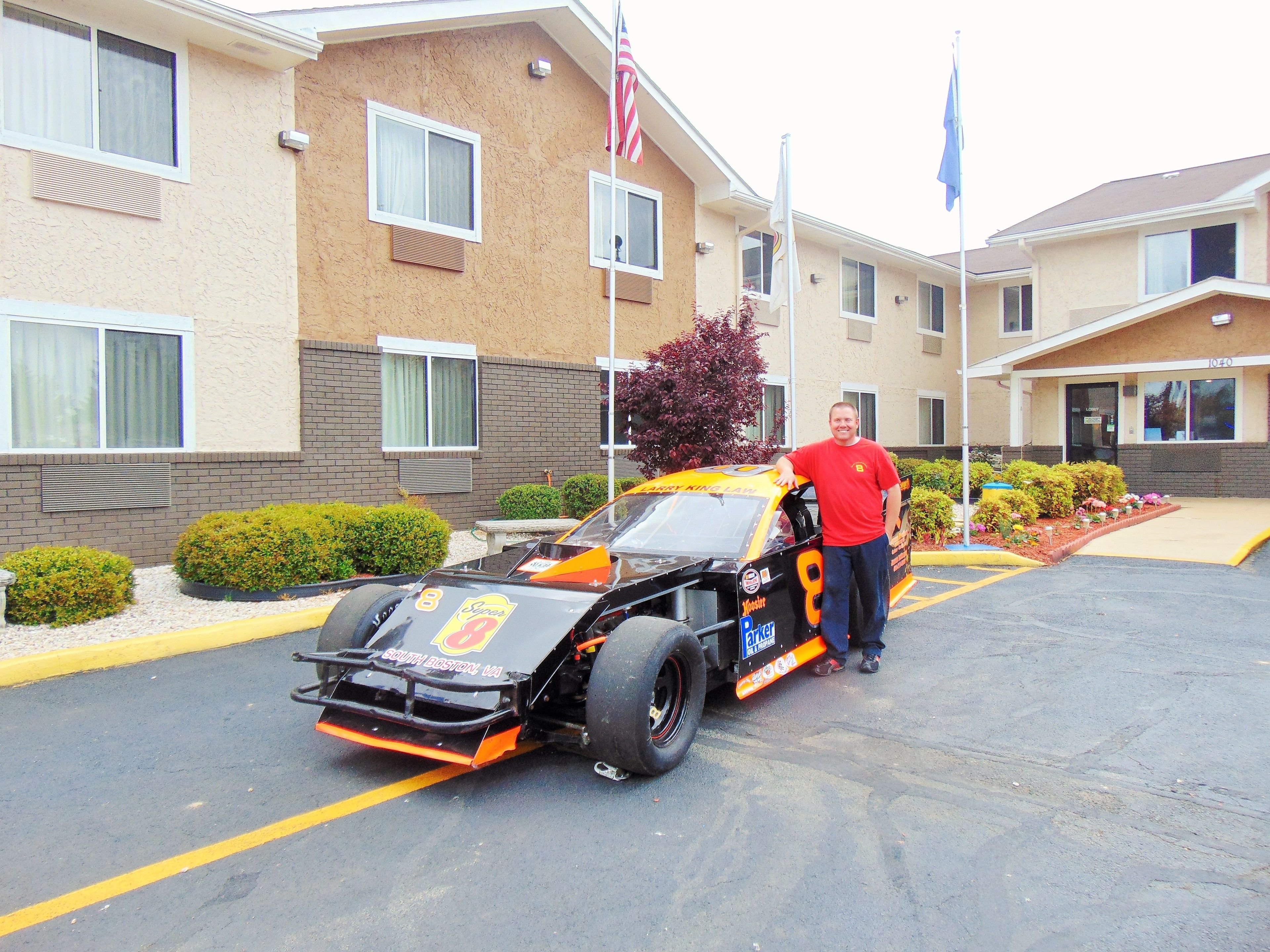 A man standing next to a race car in a parking lot with a building in the background.
